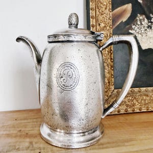 May include: A silver-colored teapot with a lid and handle. The teapot has a spout and a decorative emblem on the front. It sits on a wooden surface. The background includes a framed painting.