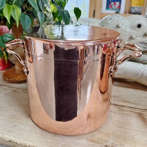 May include: A copper-colored pot with a lid and two curved handles. The pot has a cylindrical shape and reflects the surrounding environment. The pot is sitting on a wooden surface.
