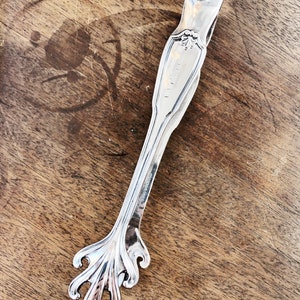 May include: A silver sugar tong with an ornate design, resting on a wooden surface. The tongs feature a decorative handle and a claw-like end for gripping sugar cubes. The word "WYNN" is visible on the handle.