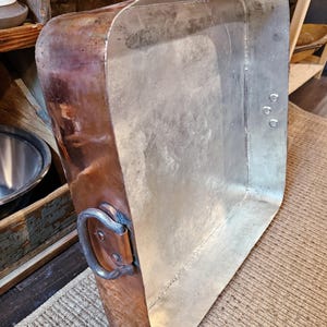 May include: A large, rectangular copper pan with a silver interior. The pan has a rounded handle and a riveted construction. The copper exterior has a rich, aged patina. The pan is likely for cooking or serving.