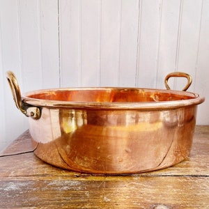 以下が含まれることがあります： 真鍮製の取っ手が2つ付いた、大きな丸い銅製の鍋。鍋は木製の台の上に置かれています。