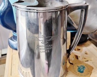 May include: A silver-toned metal pitcher featuring a prominent eagle emblem and the engraved text AMERICAN PRESIDENT LINES. The vessel has a cylindrical body, a sturdy handle, and a small pouring spout, with a small tag attached to the handle.