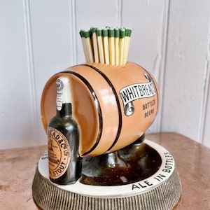 May include: A ceramic match holder shaped like a beer barrel with a bottle of Whitbread & Co. Ltd. Stout on the side. The barrel has the text "Whitbread Bottled Beers" and the base says "Ale in Bottles".