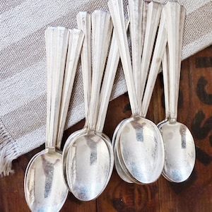 May include: A set of seven silver-toned metal spoons with a polished finish. Each spoon features a long, slender handle with vertical ribbed detailing and a rounded bowl. The spoons are arranged in a cluster, showing their reflective surfaces.