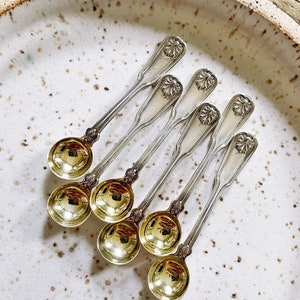 Pode incluir: Seis colheres de chá de prata com cabos ornamentados e tigelas douradas dispostas em um prato branco salpicado.