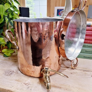 May include: A copper beverage dispenser with a tap and two handles. The dispenser has a matching copper lid with a handle. The dispenser is on a wooden surface.