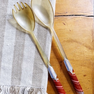 May include: A pair of serving utensils, a fork and spoon, with translucent amber-colored heads and decorative handles. The handles feature silver-colored metal accents and red and white striped agate-like material. The utensils rest on a striped linen towel.