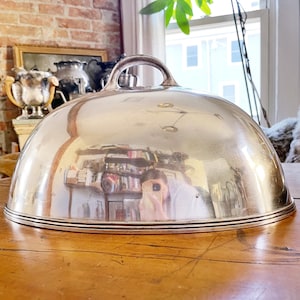May include: A silver serving dome with a handle on top. The dome is sitting on a wooden table.