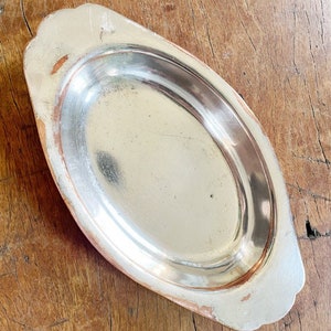 May include: A silver oval-shaped serving dish with a slightly raised rim and a smooth, polished surface. The dish has a slightly rounded edge and two small handles on either side.