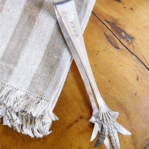 May include: Silver-toned sugar tongs with a bird claw design at the gripping end. The tongs rest on a striped, fringed linen cloth and a wooden surface. The tongs have the initials "W.R.R." engraved on one arm.
