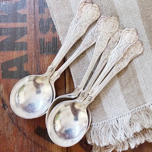 May include: A set of four silver-toned metal spoons featuring intricate embossed floral and scrollwork patterns on the handles. The spoons have rounded bowls and elongated, decorative stems that taper toward the top, showcasing a polished finish.