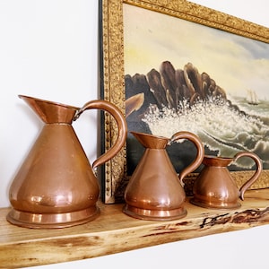 May include: A set of three graduated copper pitchers with smooth, polished surfaces, wide bases, and curved handles. Each pitcher features a tapered body and a distinct pouring spout, arranged in a row from largest to smallest in size.