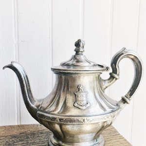 May include: A silver-toned metal teapot featuring a curved spout, a handle, and a lid topped with a small finial. The body of the teapot is decorated with vertical fluting and a central embossed crest depicting a small animal figure.