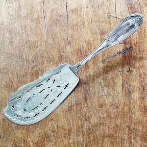 May include: A silver cake server with an ornate design and a decorative handle. The server has a scalloped edge and a pierced design.