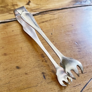 May include: Silver sugar tongs with a decorative handle and three prongs on each end. The tongs are lying on a wooden surface.
