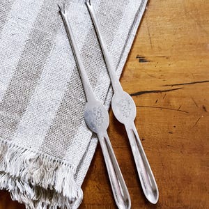 May include: Two silver cocktail forks with decorative oval handles and two prongs, resting on a fringed, striped linen napkin. The forks are on a wooden surface.