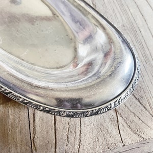 May include: A silver oval serving tray with a decorative, ornate border. The tray is made of metal and has a shiny, reflective surface.
