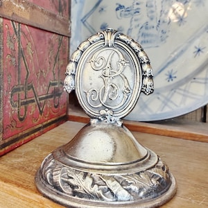 May include: Silver-toned decorative stand with an oval emblem featuring an ornate letter "B". The stand has a detailed base with leaf patterns and a flared pedestal. A red and gold box and a blue and white plate are in the background.