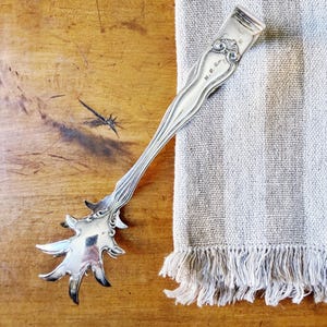 May include: A silver-colored serving utensil with an ornate handle and a starburst-shaped head. The handle has decorative flourishes and the initials "N.P.R." The utensil rests on a wooden surface next to a fringed, striped cloth.
