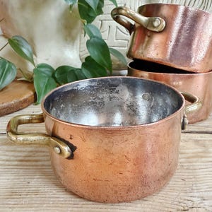 May include: A collection of antique copper pots with brass handles. The pots are various sizes and show signs of age and use. The copper has a warm, reddish-brown hue, and the handles are a shiny gold color. The pots are stacked on a wooden surface.