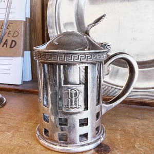 May include: A silver-colored, antique creamer with a handle and a lid. The creamer has a rectangular body with a decorative pattern and a crest. A silver plate is in the background. The creamer is on a wooden surface.