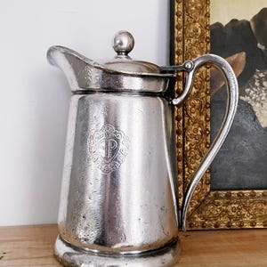May include: A silver-colored pitcher with a lid and handle. The pitcher has a spout and a decorative emblem on the front. The pitcher is sitting on a wooden surface. A painting in a gold frame is in the background.