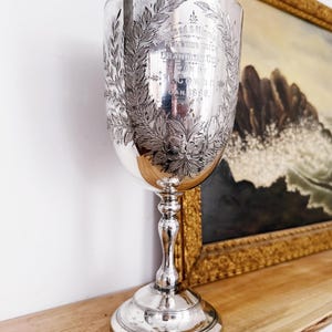 Puede incluir: Una copa de trofeo plateada con un cuenco ancho, un tallo delgado y una base circular. La copa está grabada con una corona de laurel y texto, incluyendo "Champion Cup" y la fecha "Jan. 1898". Se encuentra sobre una estantería de madera, con una pintura enmarcada al fondo.