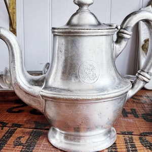 May include: A silver-toned metal teapot with a curved spout, a handle, and a lid. The body of the teapot features a circular embossed emblem depicting a figure on horseback. The surface has a brushed, matte finish with visible signs of wear.