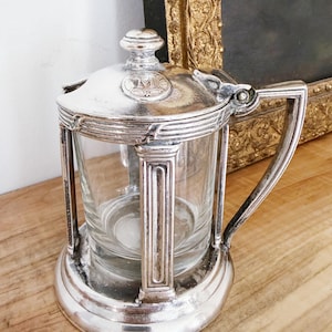 May include: A vintage silver-plated sugar bowl with a clear glass insert. The bowl has a hinged lid, a handle, and decorative detailing. The piece is set on a wooden surface, suggesting a classic or antique aesthetic.