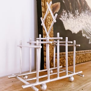 May include: A silver-colored metal napkin holder with a geometric design. The holder features a rectangular base with rounded feet and vertical bars. A diamond-shaped accent sits atop the structure. The holder is displayed on a wooden surface.