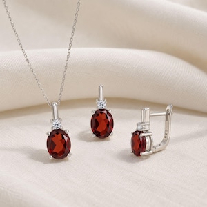 Op de afbeelding: Een zilveren ketting en een paar bijpassende oorbellen, elk voorzien van een ovaal geslepen dieprode granaat edelsteen geaccentueerd door een kleine heldere steen. De sieraden zijn gemaakt van 925 sterling zilver met een 925 keurmerk.