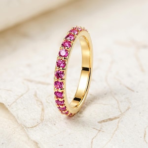 May include: A gold band ring featuring a continuous row of round, vibrant pink gemstones set along the outer edge. The metal has a polished, reflective finish, and the stones are arranged in a uniform, circular pattern around the band.