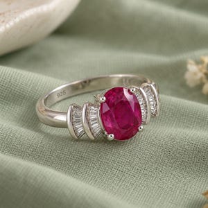 May include: A silver ring featuring a large oval-cut vibrant red gemstone at the center, flanked by three small baguette-cut clear stones on each side. The inner band is clearly stamped with the number 925, indicating sterling silver construction.