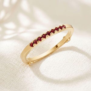 Könnte beinhalten: Ein dünner Goldring mit einer Reihe von neun kleinen, runden, tiefroten Granat-Edelsteinen, die entlang des oberen Bogens gefasst sind. Das Metall hat eine polierte Oberfläche und der Ring enthält zwei weitere winzige klare Steine an den Seiten.