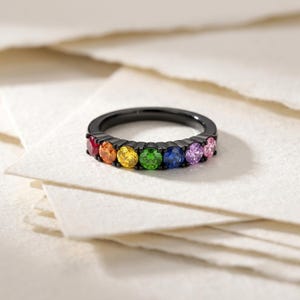 Puede incluir: Un anillo de metal negro con una hilera de siete piedras preciosas de corte redondo en un espectro de colores que incluye rojo, naranja, amarillo, verde, azul, morado y rosa, engastadas con garras en la parte frontal de la banda.
