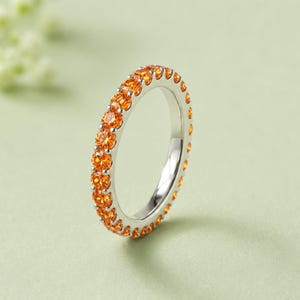 Puede incluir: Un anillo de tipo alianza en tono plateado con una hilera continua de piedras preciosas redondas de color naranja vibrante, engastadas en un canal de metal pulido. El anillo presenta un diseño circular uniforme con piedras brillantes.