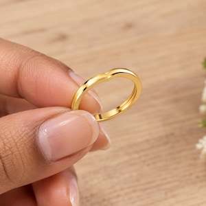 May include: A polished gold band ring featuring a subtle V-shaped chevron contour. The ring has a smooth, rounded surface and a continuous metallic finish, showcasing a simple and clean design that follows a gentle arc along the top edge.