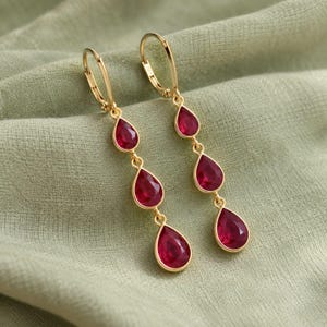 Pode incluir: Um par de brincos pendentes em tom dourado com três pedras vermelhas facetadas em forma de gota, dispostas verticalmente num design linear. Cada brinco possui um fecho de alavanca articulado e um acabamento em metal polido.
