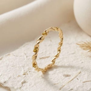 以下が含まれることがあります： ロープのようなねじれたバンドデザインが特徴の細いゴールドリング。ねじれた金属の表面に沿って、3つの小さな丸い黄色の宝石が一定の間隔で配置されており、繊細で連続した円形の構造を作り出しています。