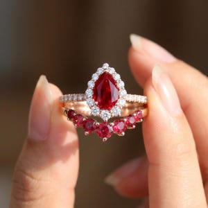 Pode incluir: Um anel de ouro rosa com uma pedra vermelha central em forma de pera rodeada por um halo de pequenas pedras brancas redondas. A banda inclui uma secção curva adornada com cinco pedras vermelhas redondas e pedras brancas adicionais.