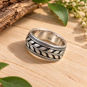 May include: A silver-toned ring featuring a central band with a repeating leaf pattern, bordered by two rows of small raised dots. The interior of the band is engraved with the number 925, indicating the metal composition of the jewelry piece.