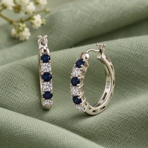 May include: A pair of sterling silver hoop earrings featuring an alternating pattern of round-cut dark blue sapphires and clear white diamonds. The inner band of the hoop is clearly stamped with the 925 hallmark indicating sterling silver composition.