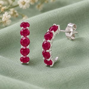 Puede incluir: Un par de pendientes de botón de plata de ley, cada uno con cinco rubíes rojos vibrantes de talla redonda en una fila vertical ligeramente curvada. El cierre de plata tiene grabado el sello 925, que indica la pureza del metal del conjunto.