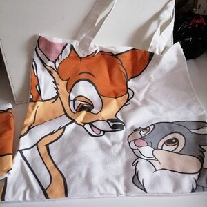 Puede incluir: Una bolsa de lona blanca con un ciervo marrón y naranja y un conejo gris impresos en ella. El ciervo mira hacia la derecha y el conejo mira hacia la izquierda. La bolsa tiene dos asas blancas.
