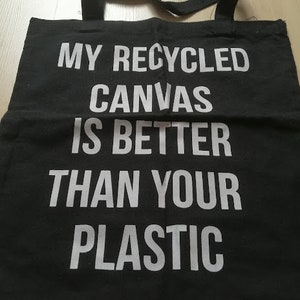 May include: Black canvas tote bag with white text: "MY RECYCLED CANVAS IS BETTER THAN YOUR PLASTIC." The bag has two black handles. Made from durable, eco-friendly material, it's a reusable shopping bag.