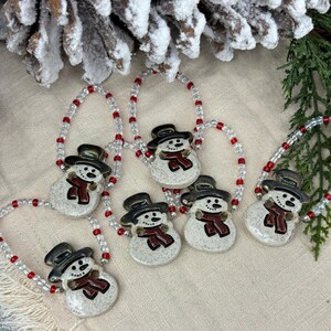 May include: Six ceramic snowman ornaments with red scarves and black hats, strung on a white and red beaded garland. The ornaments are on a beige fabric surface.