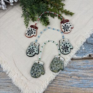 May include: Five ceramic mitten ornaments with snowflake designs, strung together with white and blue beads. The mittens are in shades of red, white, black, and green.