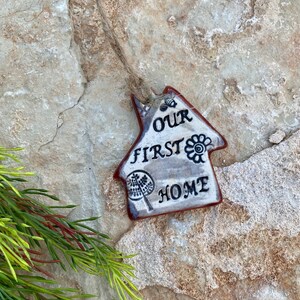 May include: A white ceramic house-shaped ornament with the words "Our First Home" in black lettering. The ornament has a brown rope attached to the top and is hanging on a stone wall.