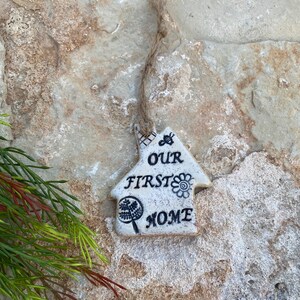 May include: A white ceramic house-shaped ornament with black lettering that reads "Our First Home". The ornament has a twine string for hanging and is decorated with a bee, a flower, and a tree.