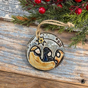 May include: A ceramic Christmas ornament with a nativity scene design. The round ornament features a star, figures, and a manger in shades of blue, brown, and gold. It has a twine loop for hanging and is set against a backdrop of greenery and red berries.
